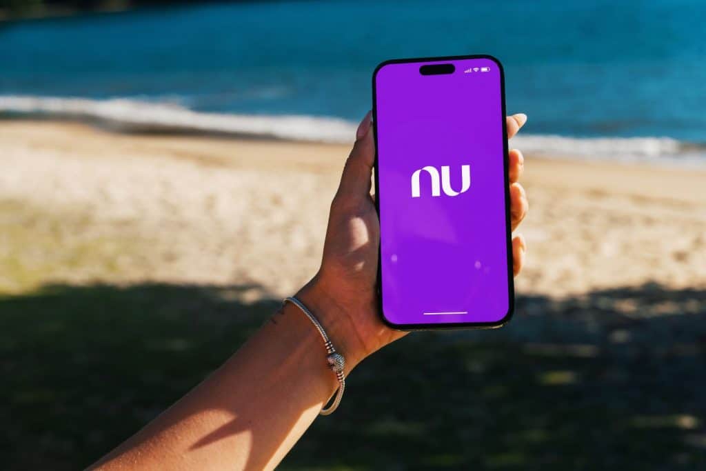 Garota na praia com smartphone iPhone 14 Pro visualizando o app do Nubank na tela, cenário à beira-mar no Rio de Janeiro.