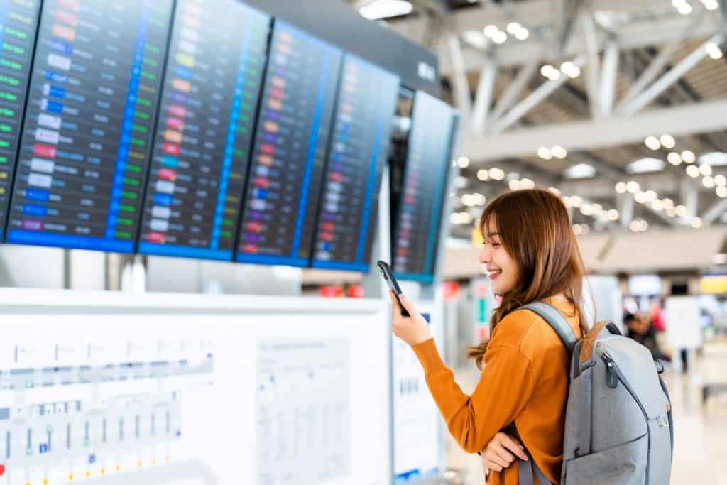 Jovem mulher asiática em aeroporto internacional, usando smartphone e checando o painel de informações do voo.