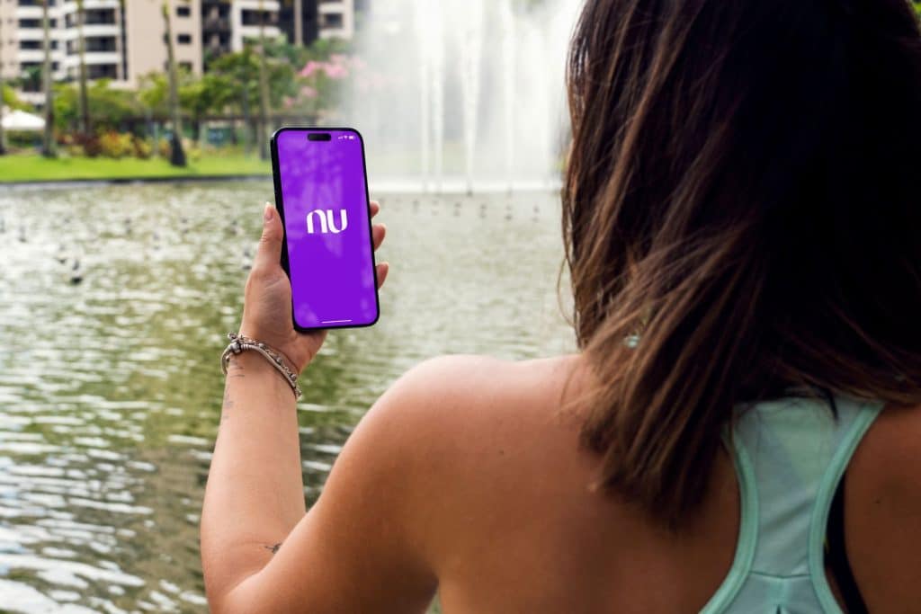 Menina no parque com smartphone iPhone 14 Pro mostrando o aplicativo do Nubank na tela, cenário de lago ao fundo em Rio de Janeiro.