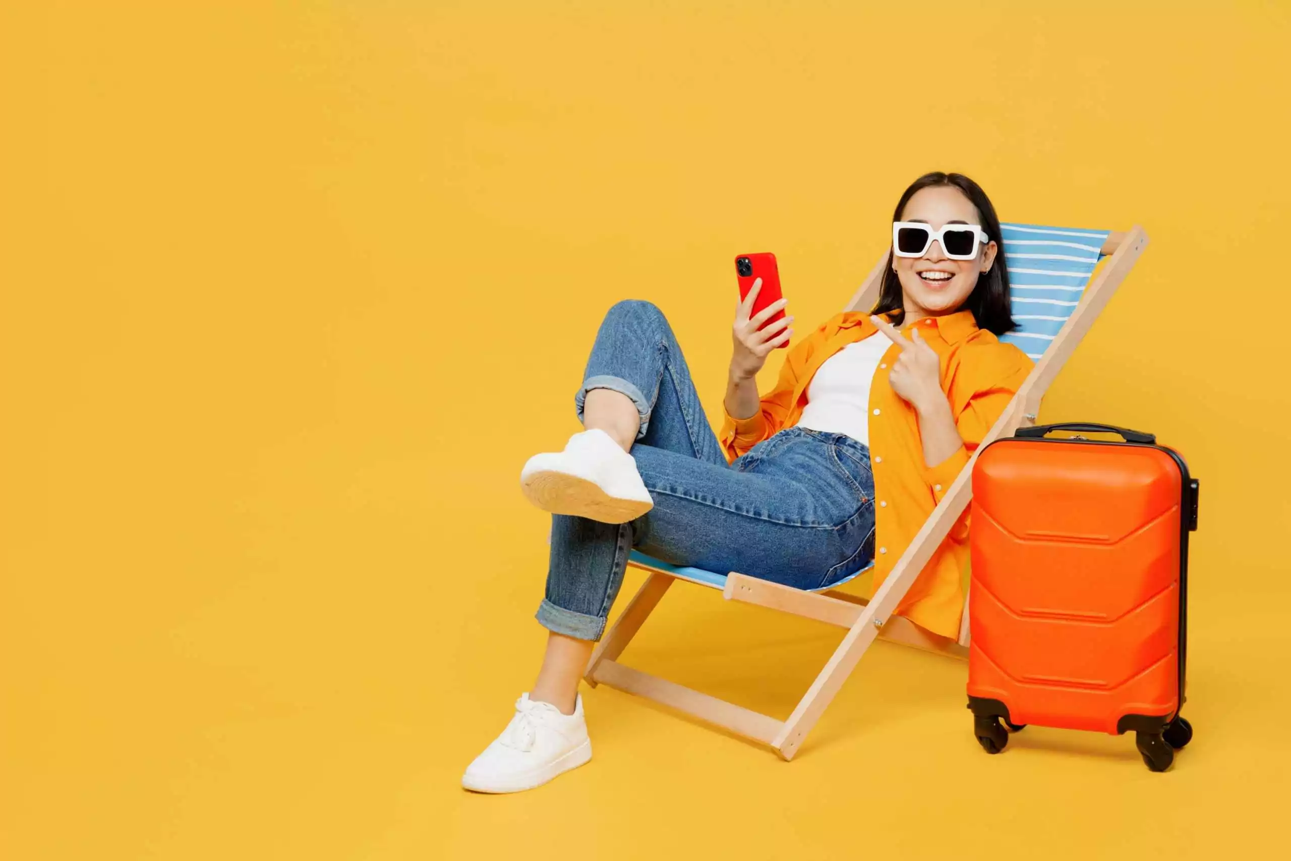 Jovem mulher vestindo roupas de verão sentada em uma espreguiçadeira usando o celular isolada em um fundo amarelo liso. Turista viajando para o exterior no tempo livre para descansar e relaxar. Conceito de viagem de avião.