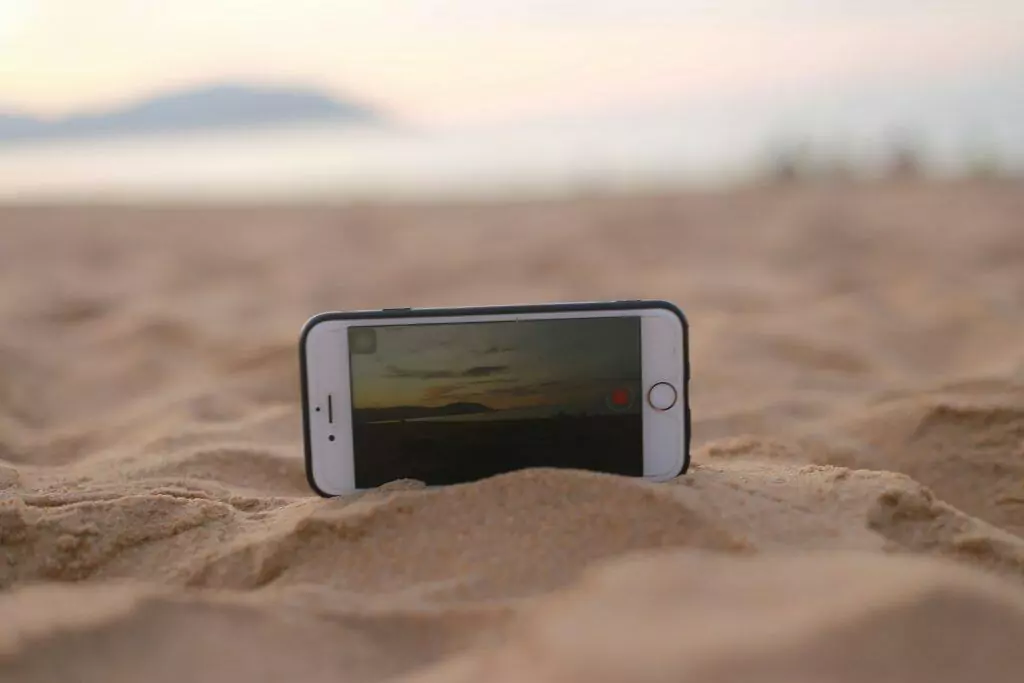 Celular à prova d'água? Entenda a diferença entre IP e ATM Guia Região dos Lagos 5 iPhone na areia de uma praia, ressaltando a importância da resistência à água