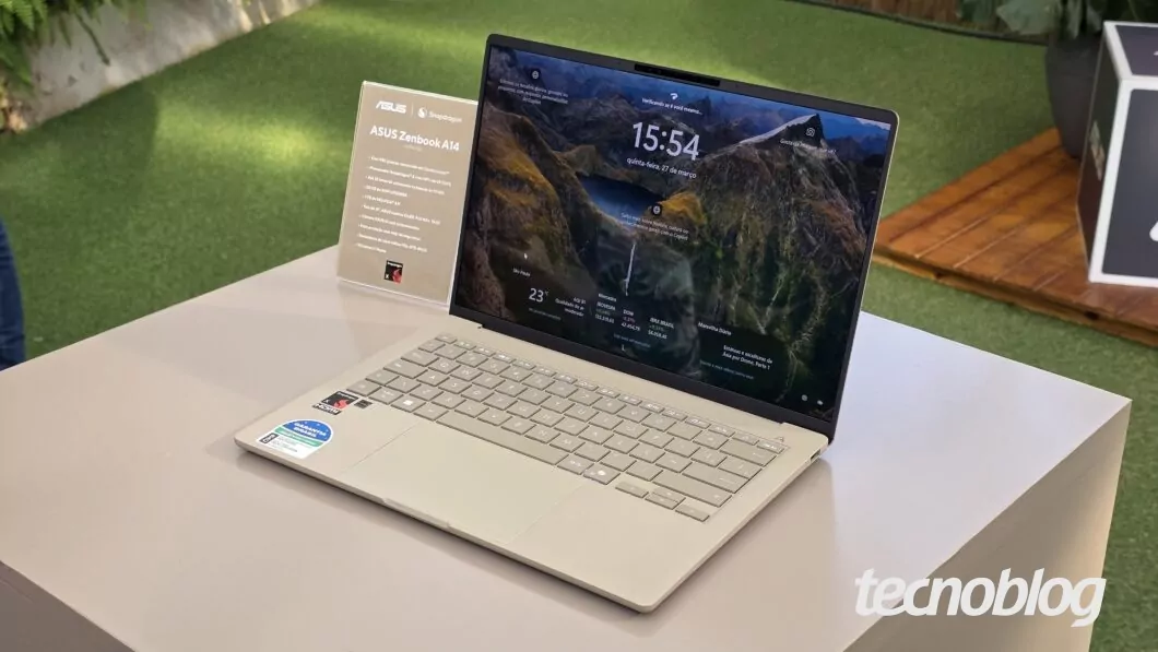 A imagem mostra um notebook ASUS Zenbook A14 exposto sobre uma mesa em um ambiente bem iluminado e decorado com grama sintética e plantas ao fundo. Ao lado do notebook, há um pequeno display informativo com o nome e algumas especificações do modelo, destacando o processador Snapdragon. A tela do notebook exibe o sistema Windows com o recurso de tela de bloqueio ativo, mostrando o horário (15:54), a data (quinta-feira, 21 de março) e informações sobre o clima e localização. O design do laptop é moderno e elegante, com acabamento metálico claro e teclado em tom semelhante. No canto inferior direito da imagem, aparece o logo do site Tecnoblog.