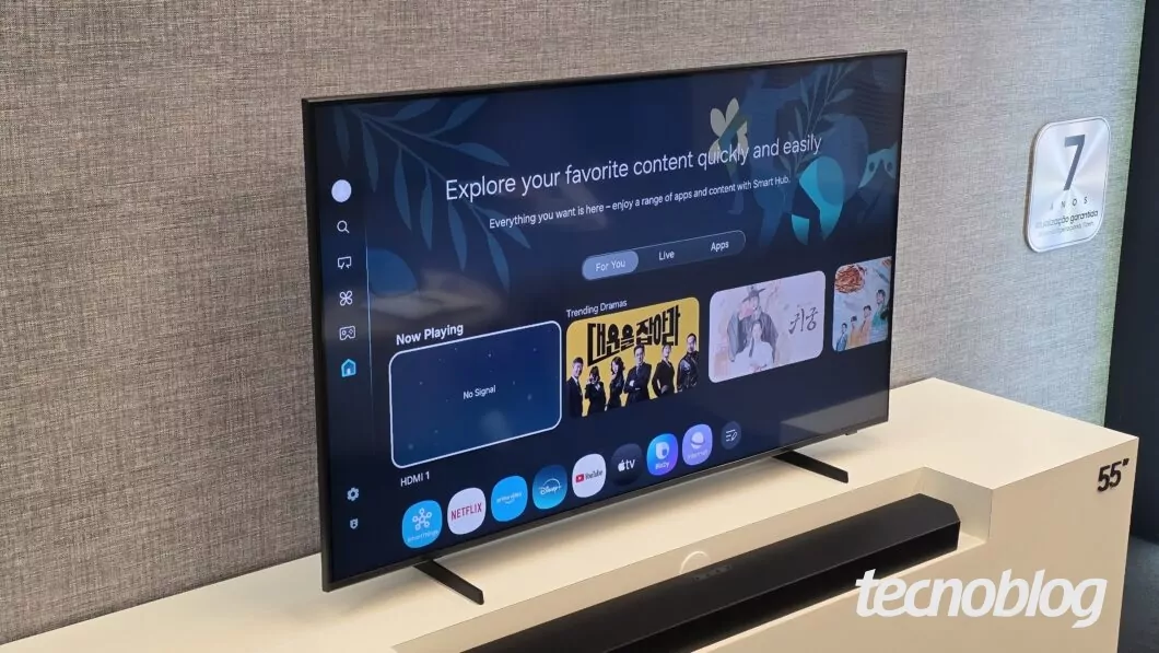 Tela de smart TV de 55 polegadas exibindo menu inicial com opções de apps como Netflix, Prime Video, Disney+, YouTube e Apple TV. No centro da tela, texto em inglês: "Explore your favorite content quickly and easily" e abaixo, seleção de dramas em destaque. A TV está sobre suporte branco e há uma soundbar preta à frente. Ao lado, placa com texto "7 anos Atualização garantida Sistema Operacional Tizen".